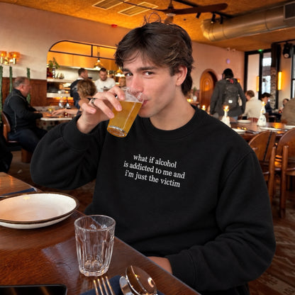 A man sitting at a restaurant table drinking from a glass, wearing a black crewneck sweatshirt with the white printed text 'what if alcohol is addicted to me and I'm just the victim'.