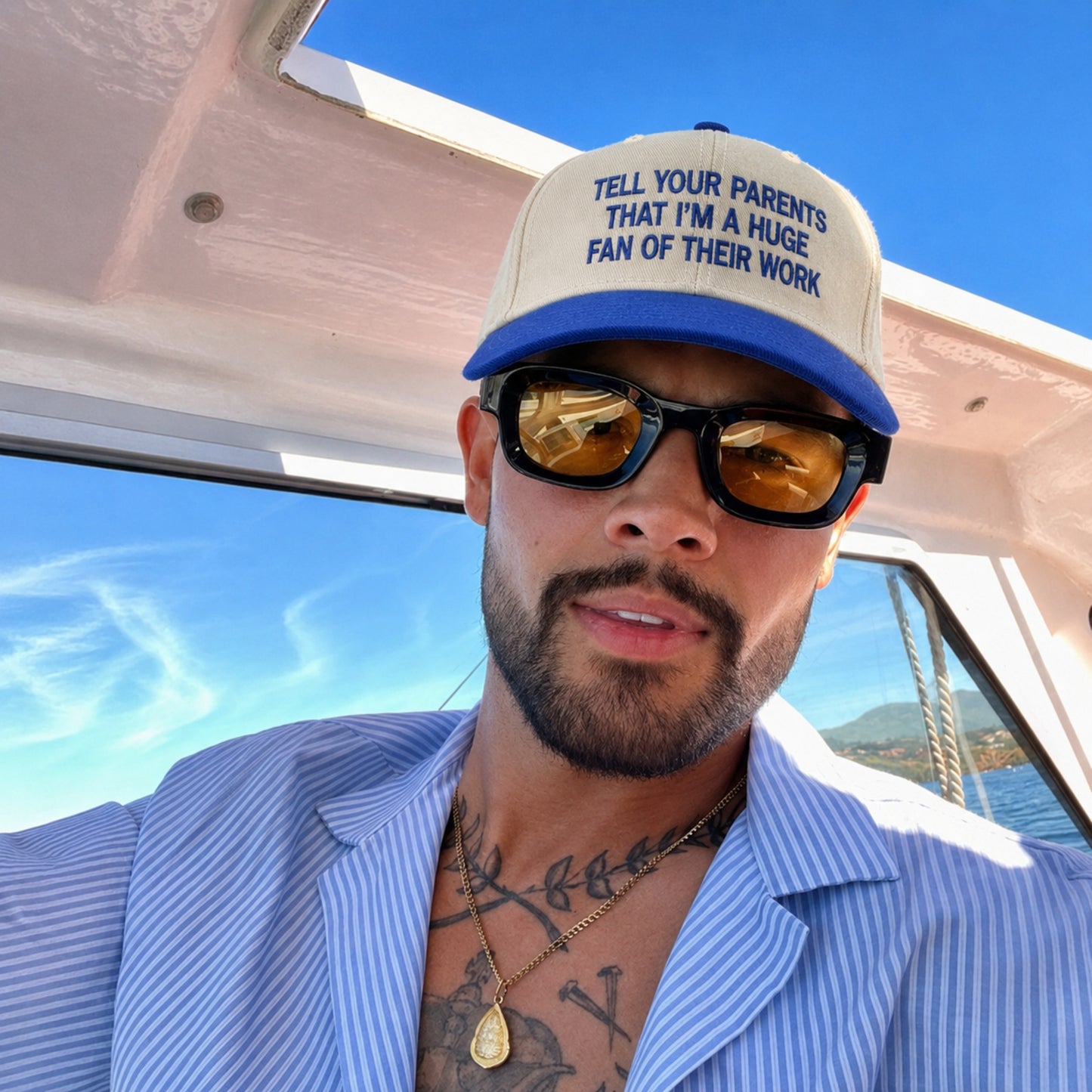 A man wearing a beige and blue trucker hat with the text 'TELL YOUR PARENTS THAT I'M A HUGE FAN OF THEIR WORK' and black sunglasses, posing on a boat under a blue sky.