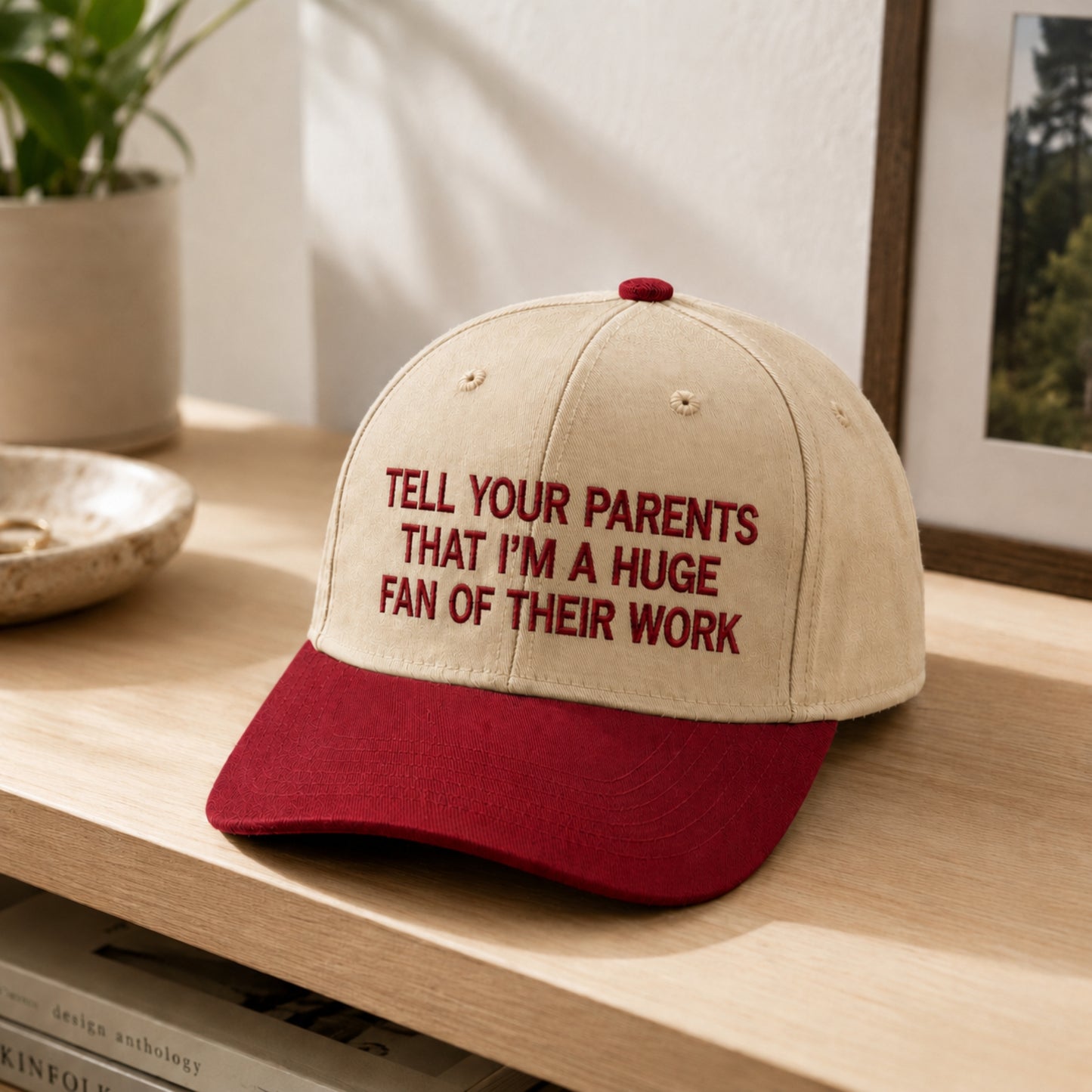 A baseball cap with a beige crown and a dark red curved brim, featuring the embroidered text 'TELL YOUR PARENTS THAT I'M A HUGE FAN OF THEIR WORK' in dark red. The cap is styled on a light wooden shelf with a potted plant and framed picture in the background.