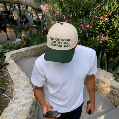 A man wearing a beige and dark green baseball cap with the text 'TELL YOUR PARENTS THAT I'M A HUGE FAN OF THEIR WORK' in green lettering. The cap is shown in an outdoor garden setting.