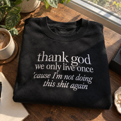 Black embroidered sweatshirt folded on a rustic wooden table by a sunny window, featuring the white quote “thank god we only live once ’cause I’m not doing this shit again,” styled with a coffee cup and green plants.