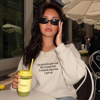 A woman sitting at an outdoor cafe table wearing a light beige graphic sweatshirt with black text, black sunglasses, and holding a yellow-topped iced coffee cup.