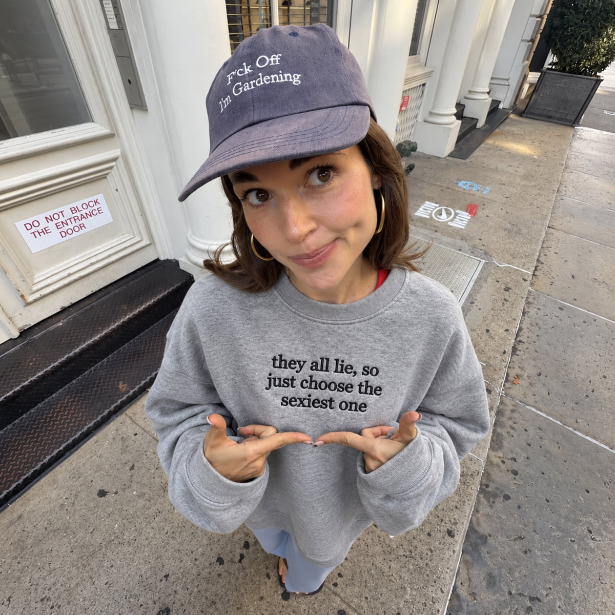 Woman wearing a sport grey embroidered sweatshirt featuring the funny quote “they all lie, so just choose the sexiest one” in black stitching, paired with a washed baseball cap, posing on a city sidewalk and pointing at the chest embroidery.