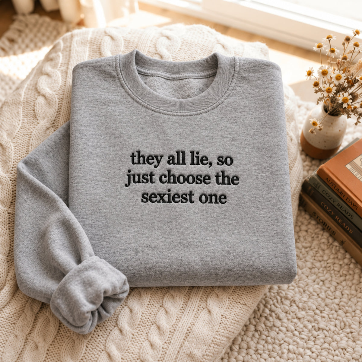 Folded sport grey embroidered sweatshirt featuring the funny quote “they all lie, so just choose the sexiest one” in black stitching, displayed on a cozy cream cable-knit blanket with warm natural sunlight, dried flowers, and vintage books.