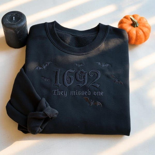 A folded black crewneck sweatshirt with a gothic-style '1692 They missed one' graphic and bat silhouettes, styled on a light surface with a black candle and a small orange pumpkin.