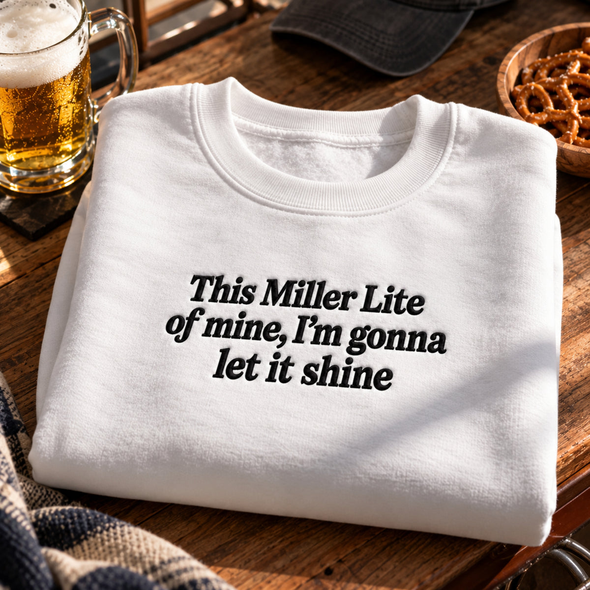 A folded white crewneck T-shirt with black text reading 'This Miller Lite of mine, I'm gonna let it shine' on a rustic wooden table, surrounded by a beer mug, a cap, and pretzels.