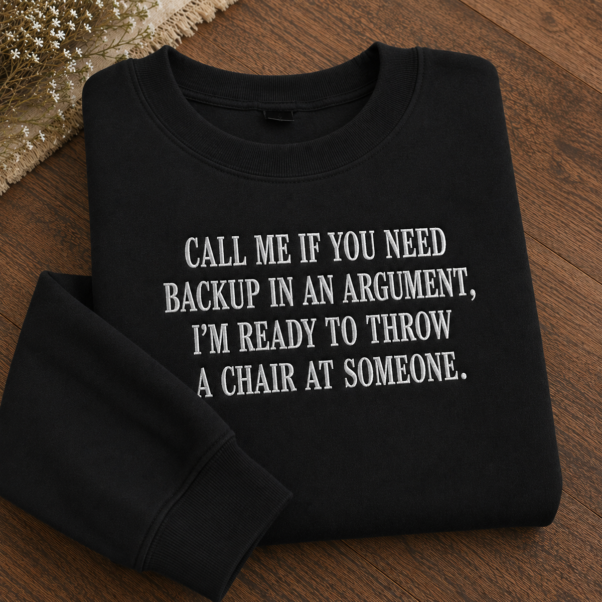 A folded black crewneck sweatshirt with white text reading 'CALL ME IF YOU NEED BACKUP IN AN ARGUMENT, I'M READY TO THROW A CHAIR AT SOMEONE.' displayed on a wooden surface with dried flowers in the corner.