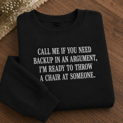 A folded black crewneck sweatshirt with white text reading 'CALL ME IF YOU NEED BACKUP IN AN ARGUMENT, I'M READY TO THROW A CHAIR AT SOMEONE.' displayed on a wooden surface with dried flowers in the corner.