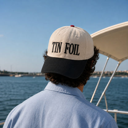 A person viewed from behind wearing a baseball cap with 'TIN FOIL' embroidered in black on an off-white crown and black brim, standing on a boat with water and sky in the background.