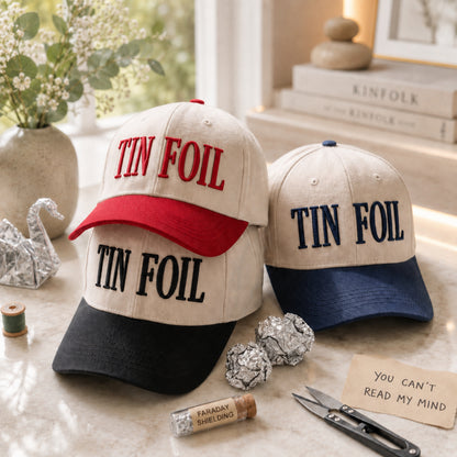 Three baseball caps with 'TIN FOIL' embroidered text are arranged on a tabletop with decorative props including foil balls, scissors, thread, a small vial, and books in the background.