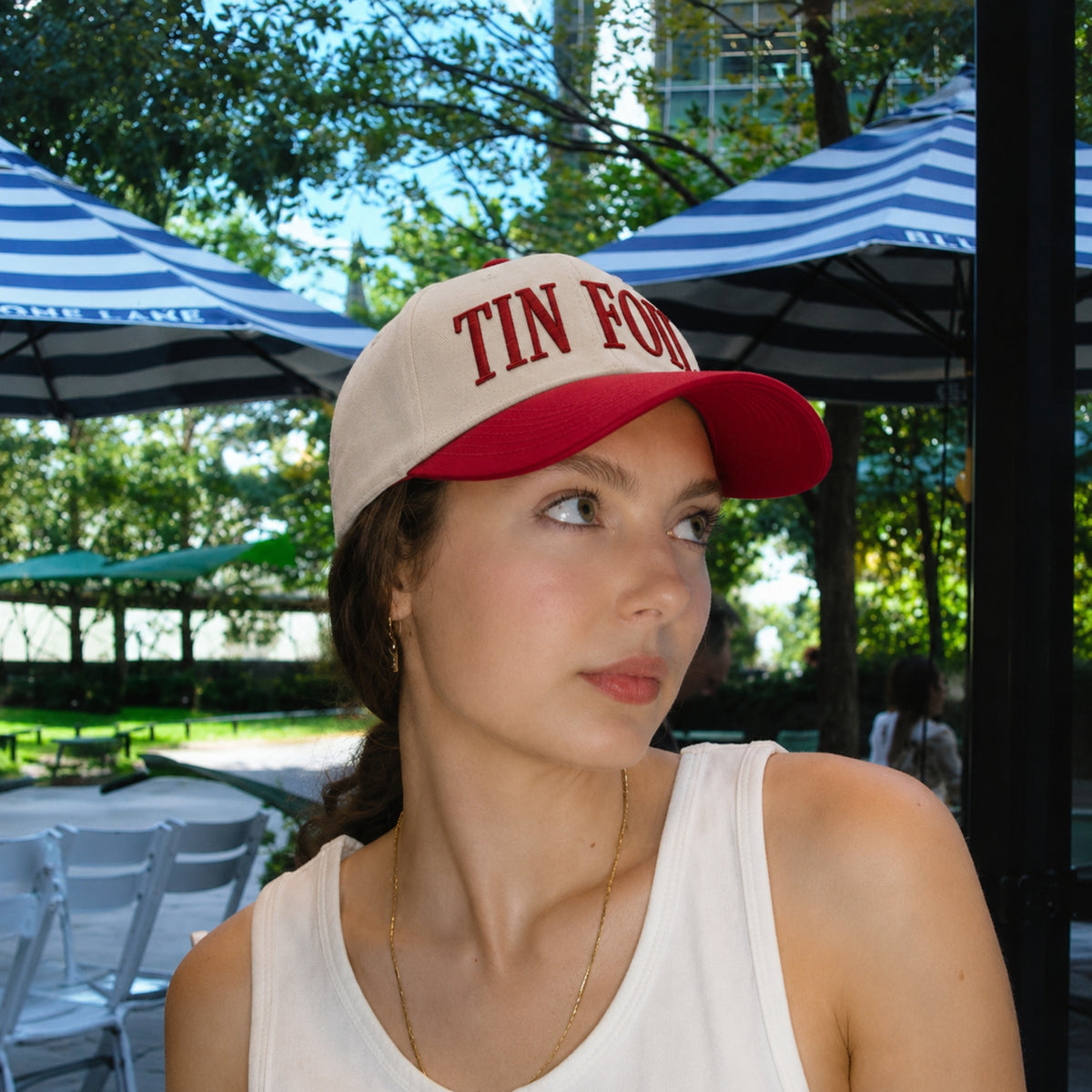 A woman wearing a beige and red baseball cap with 'TIN FORT' embroidered in red on the front, photographed outdoors under blue-and-white striped patio umbrellas.