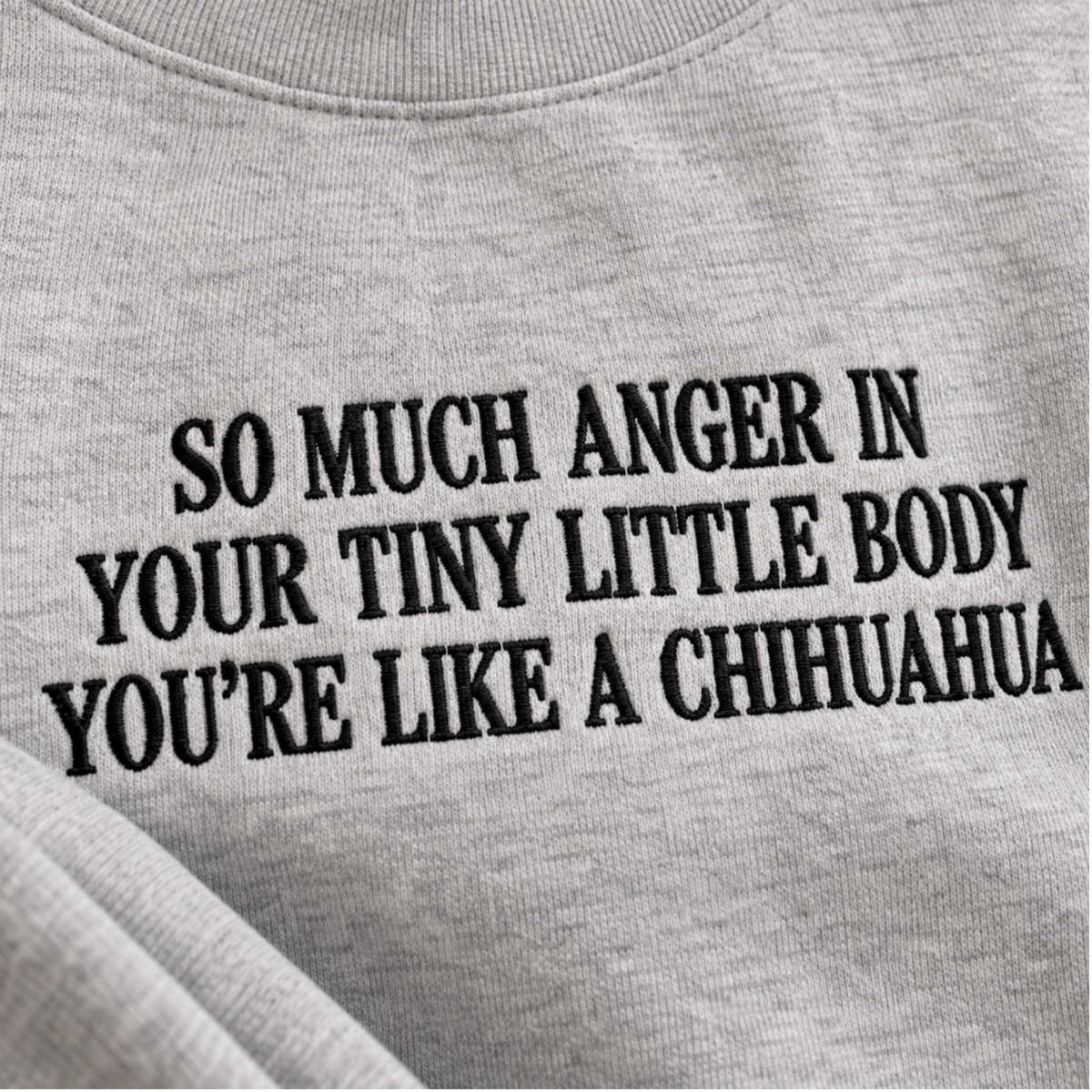 Close-up of a light gray heather crewneck sweatshirt with black embroidered text that reads 'SO MUCH ANGER IN YOUR TINY LITTLE BODY YOU'RE LIKE A CHIHUAHUA'.