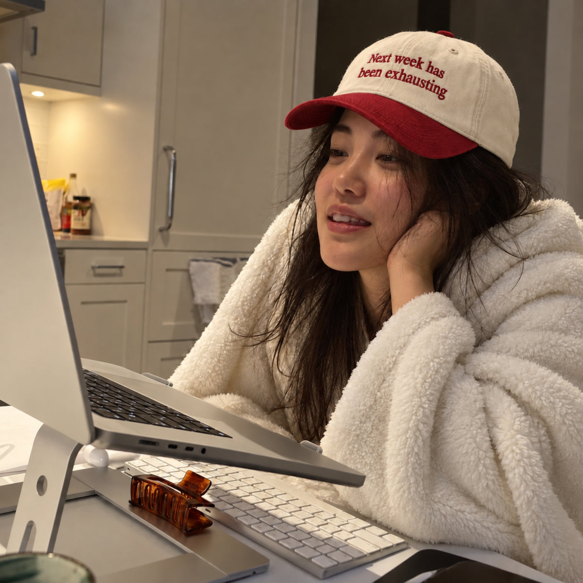 Woman wearing a cream and red embroidered baseball cap with “Next week has been exhausting” text, sitting at a desk and looking at a laptop in a cozy home setting.