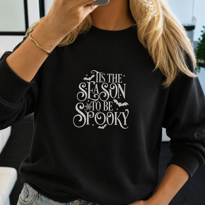 A woman wearing a black crewneck sweatshirt with a white graphic print that reads 'IT IS THE SEASON TO BE SPOOKY' with bats and a spider design. She is holding a phone in front of her face.