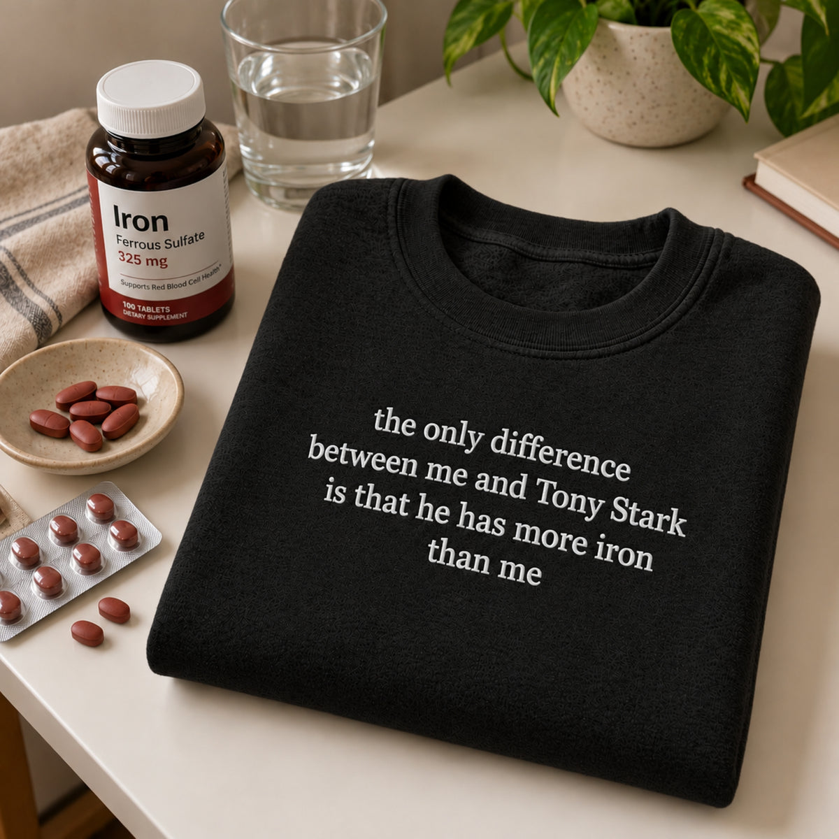 A folded black crewneck T-shirt with white text reading 'the only difference between me and Tony Stark is that he has more iron than me' sits on a tabletop surrounded by pill bottles, loose red tablets, a glass of water, and a plant.