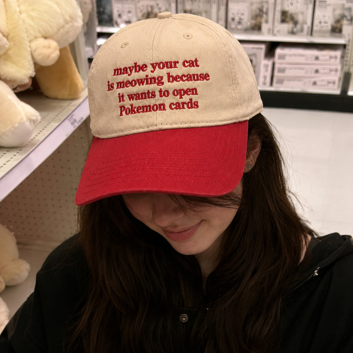 A person wearing a beige and red baseball cap with red embroidered text that reads 'maybe your cat is meowing because it wants to open Pokemon cards', photographed in a retail store setting.