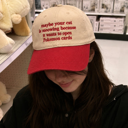 A person wearing a beige and red baseball cap with red embroidered text that reads 'maybe your cat is meowing because it wants to open Pokemon cards', photographed in a retail store setting.