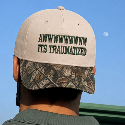A man viewed from behind wearing a beige baseball cap with dark green embroidered text reading 'AWWWWWWWWW ITS TRAUMATIZED' and a camouflage-patterned visor, set against a blue sky.