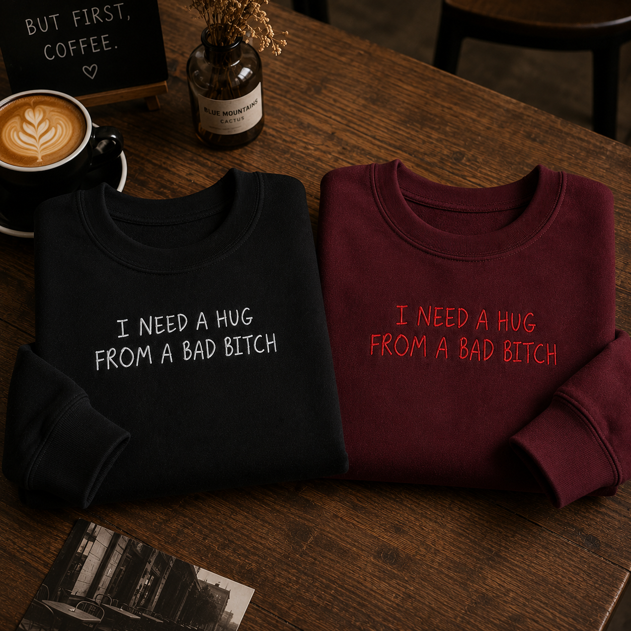 Two folded sweatshirts on a wooden table, one black with white text reading 'I NEED A HUG FROM A BAD BITCH' and one burgundy with red text reading the same phrase. A coffee cup, small bottle, and sign are visible in the background.