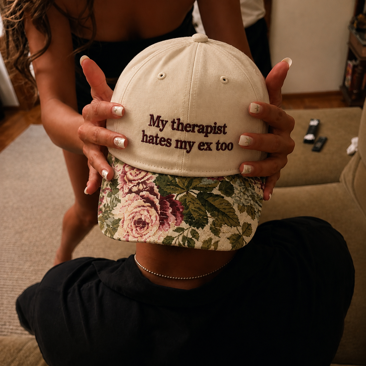Two hands holding up a beige baseball cap embroidered with "My therapist hates my ex too" in brown text and featuring a vintage floral printed brim.