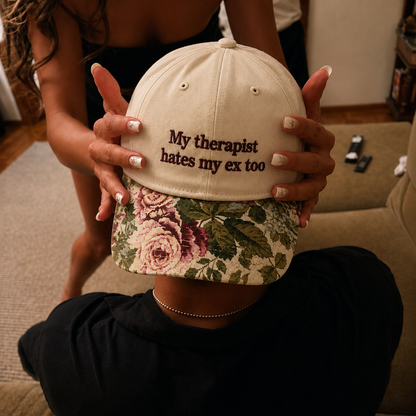 Two hands holding up a beige baseball cap embroidered with "My therapist hates my ex too" in brown text and featuring a vintage floral printed brim.