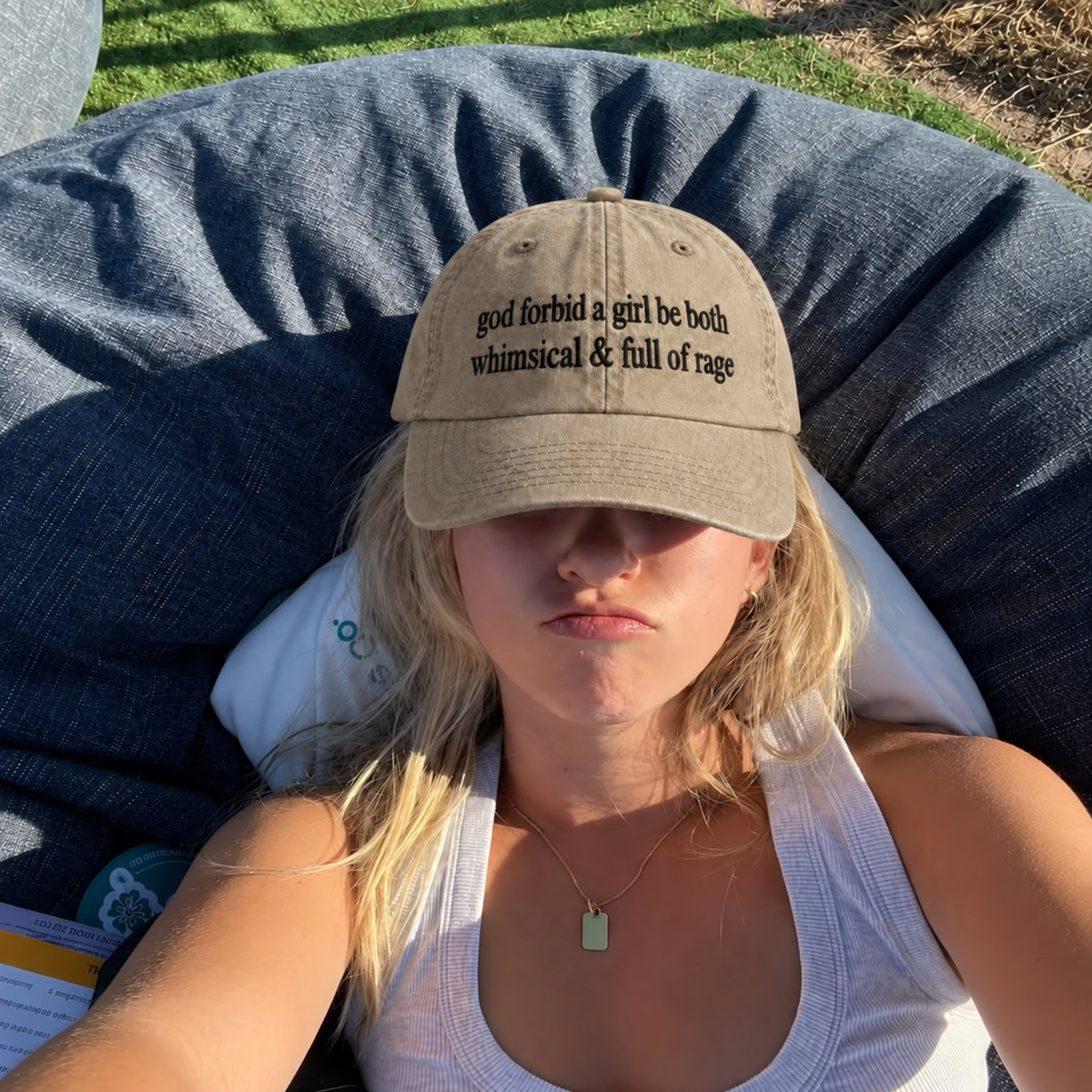 A woman reclining outdoors on a dark blue bean bag chair, wearing a beige baseball cap with black embroidered text that reads 'god forbid a girl be both whimsical & full of rage'.
