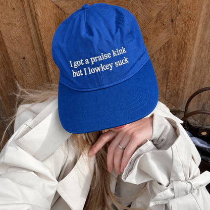 A person wearing a bright royal blue baseball cap with white embroidered text that reads 'I got a praise kink but I lowkey suck', posed in front of a rustic wooden wall.