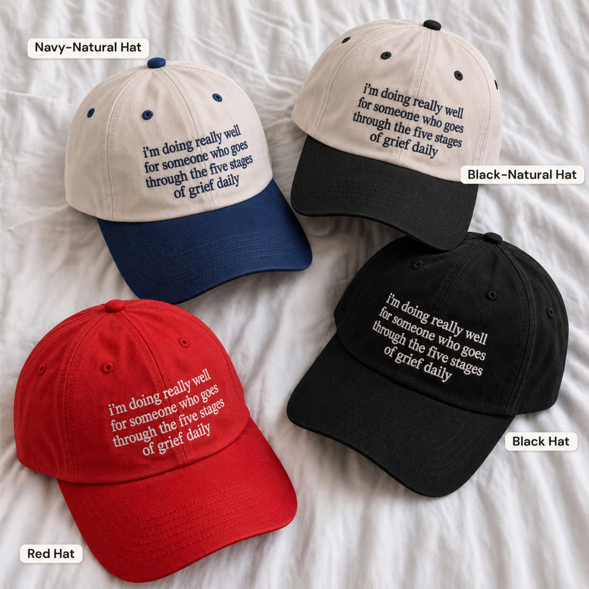 Four baseball caps arranged on white fabric, each with embroidered text reading 'I'm doing really well for someone who goes through the five stages of grief daily.' The hats are shown in four colorways labeled in the image: Navy-Natural Hat, Black-Natural Hat, Red Hat, and Black Hat.