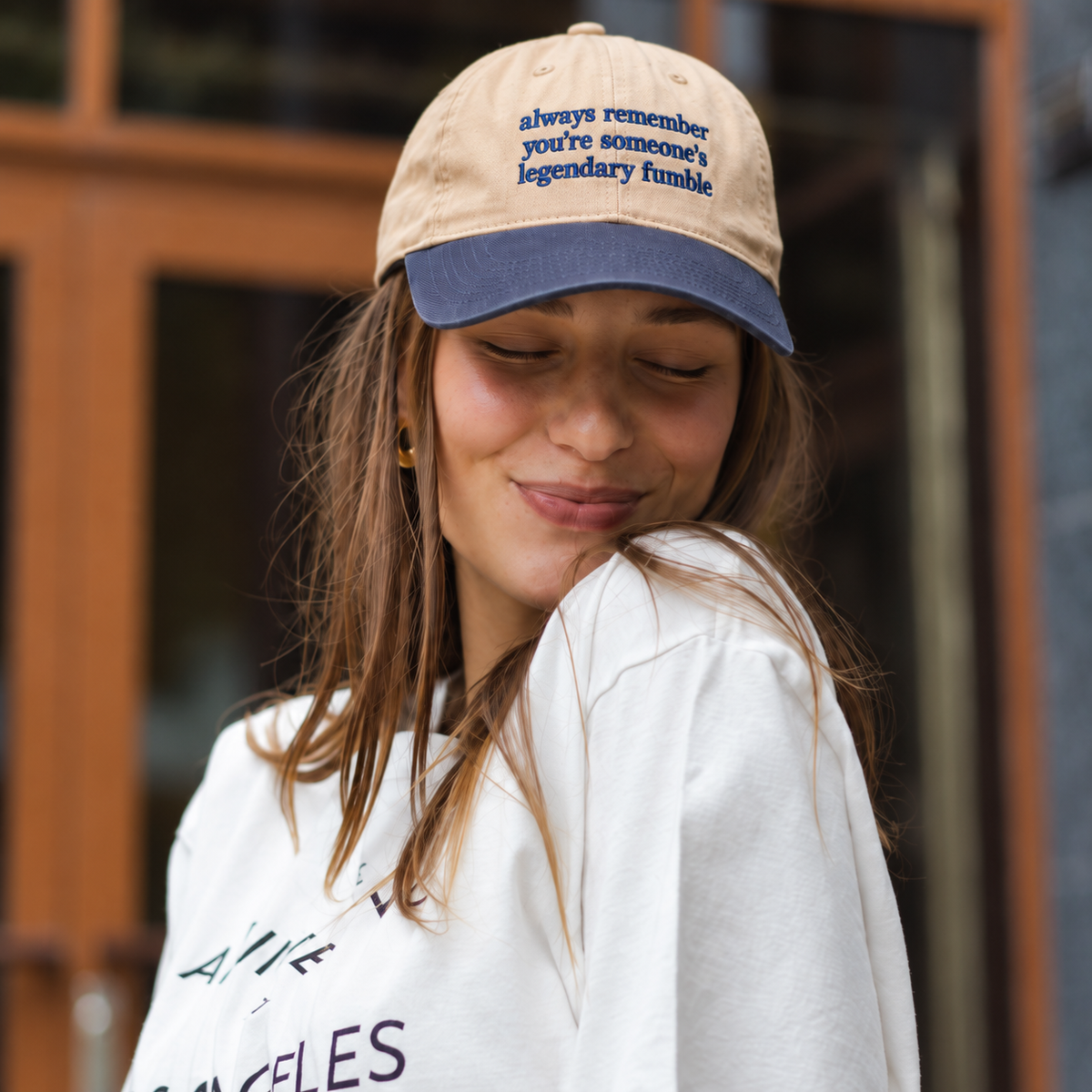 A woman wearing a beige and navy blue baseball cap with embroidered text that reads 'always remember you're someone's legendary fumble'.