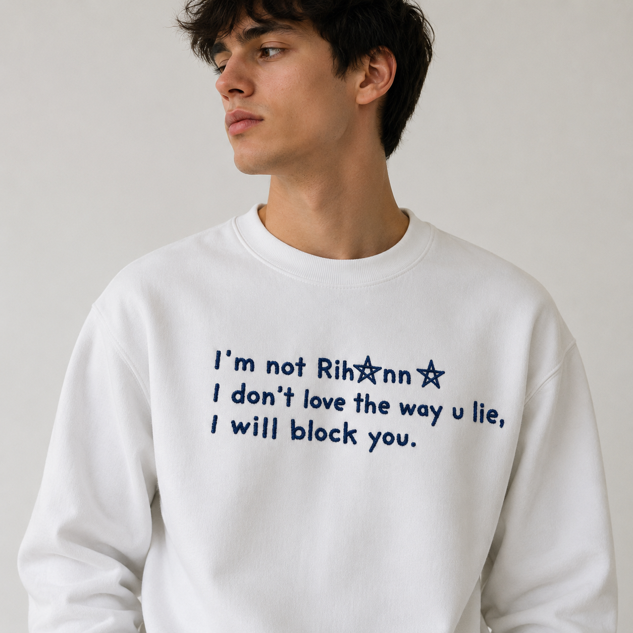 Young man with dark hair wearing a white crewneck sweatshirt embroidered with black text reading "I'm not Rihanna I don't love the way u lie, I will block you."