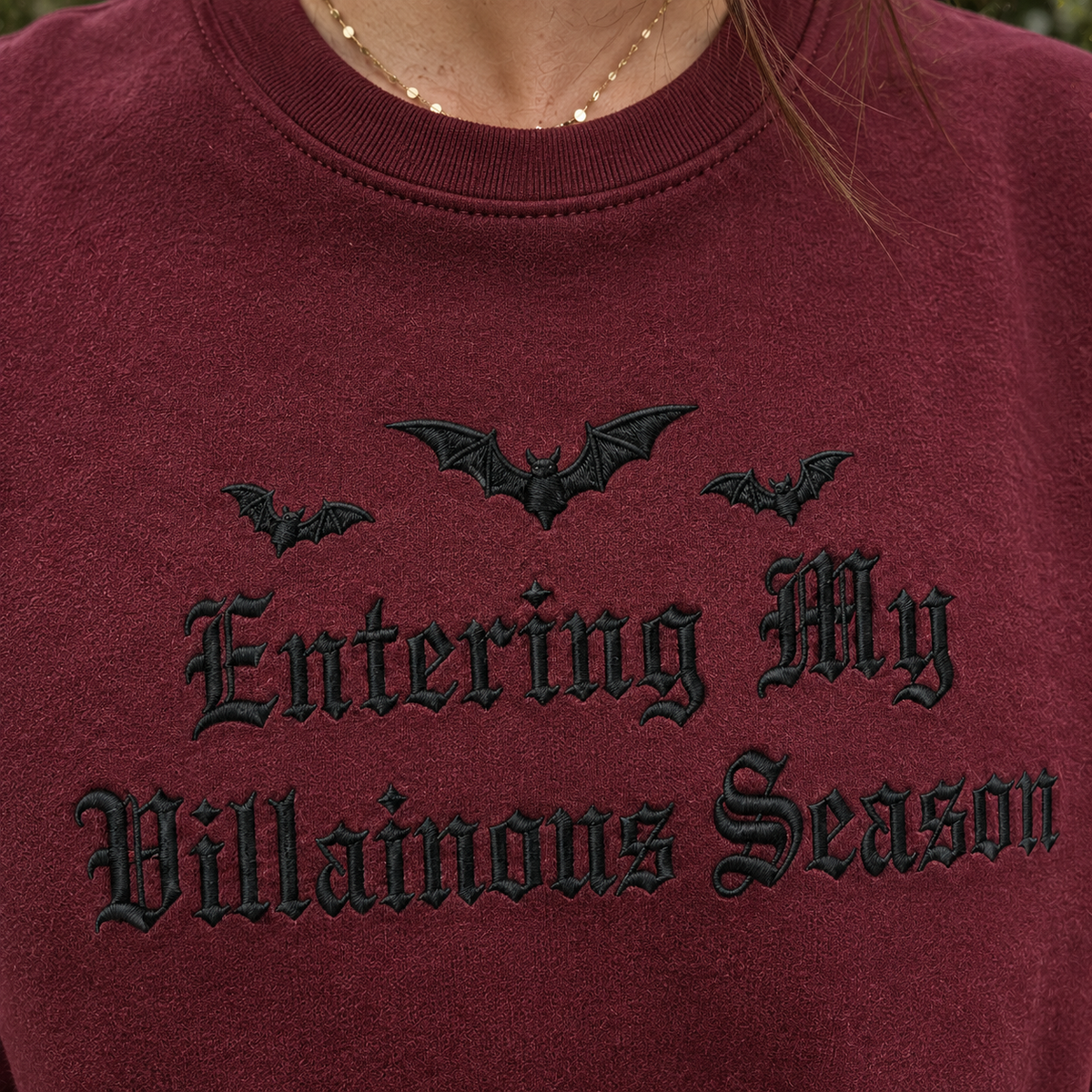 A close-up of a person wearing a burgundy crewneck sweatshirt with black embroidered gothic text reading 'Entering My Villainous Season' and three black bat silhouettes above it.