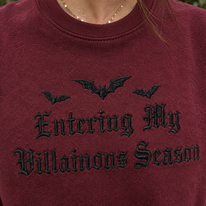 A close-up of a person wearing a burgundy crewneck sweatshirt with black embroidered gothic text reading 'Entering My Villainous Season' and three black bat silhouettes above it.