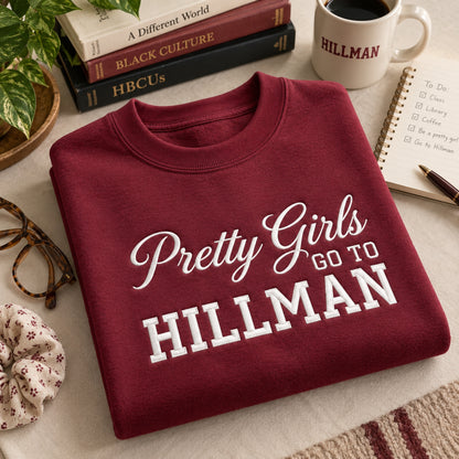 A folded maroon crewneck sweatshirt with white text reading 'Pretty Girls GO TO HILLMAN' is displayed on a light fabric surface. The scene includes books, a coffee mug with 'HILLMAN' printed on it, eyeglasses, a notebook, and a small plant as styled props.