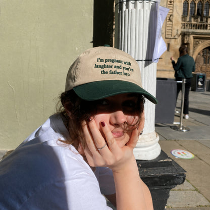 A woman wearing a beige and dark green baseball cap with the text 'I'm pregnant with laughter and you're the father bro' resting her head in her hands outdoors.