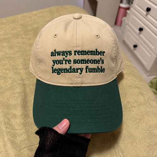 A beige and dark green baseball cap held by a hand, featuring the embroidered text 'always remember you're someone's legendary fumble' on the front panel.