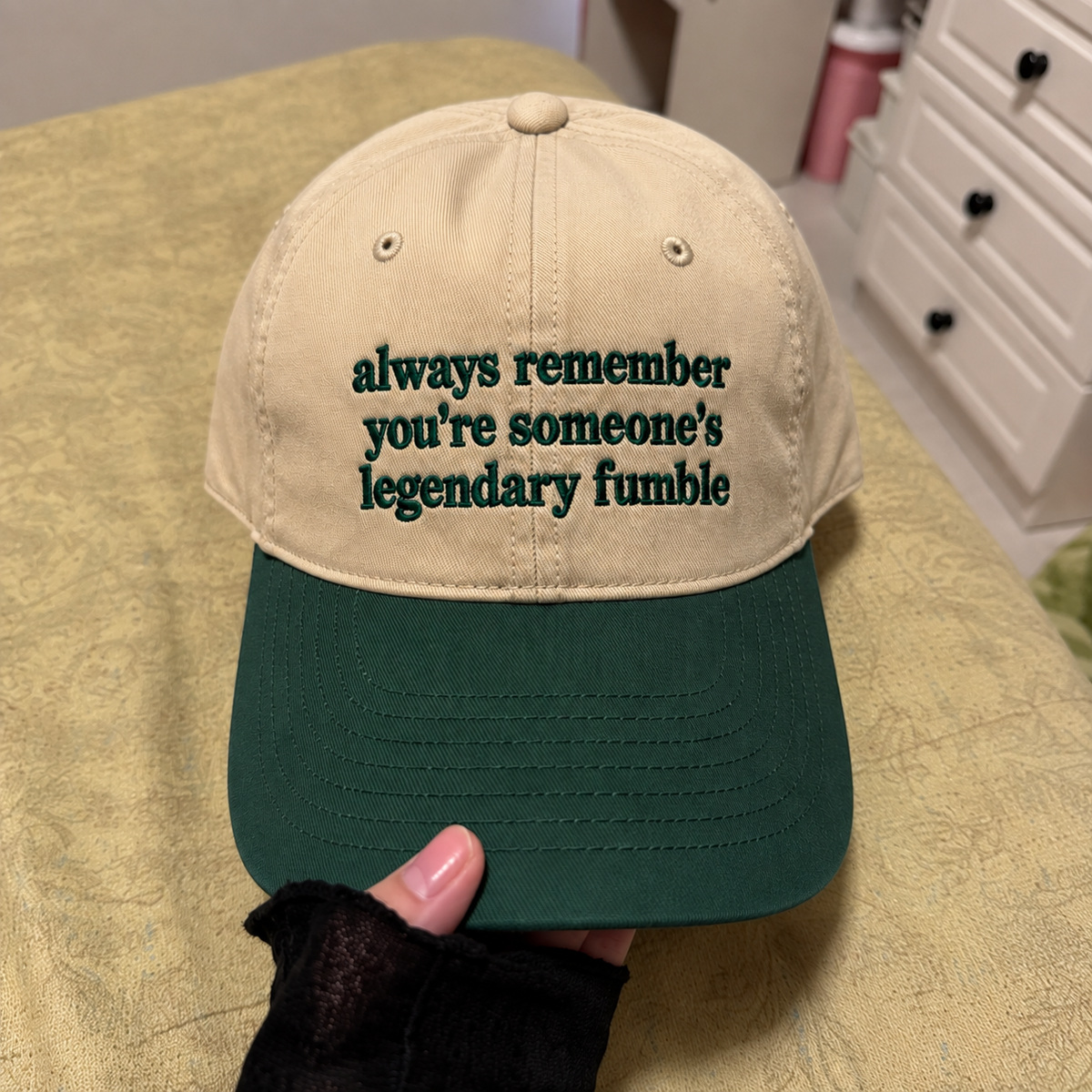 A beige and dark green baseball cap held by a hand, featuring the embroidered text 'always remember you're someone's legendary fumble' on the front panel.