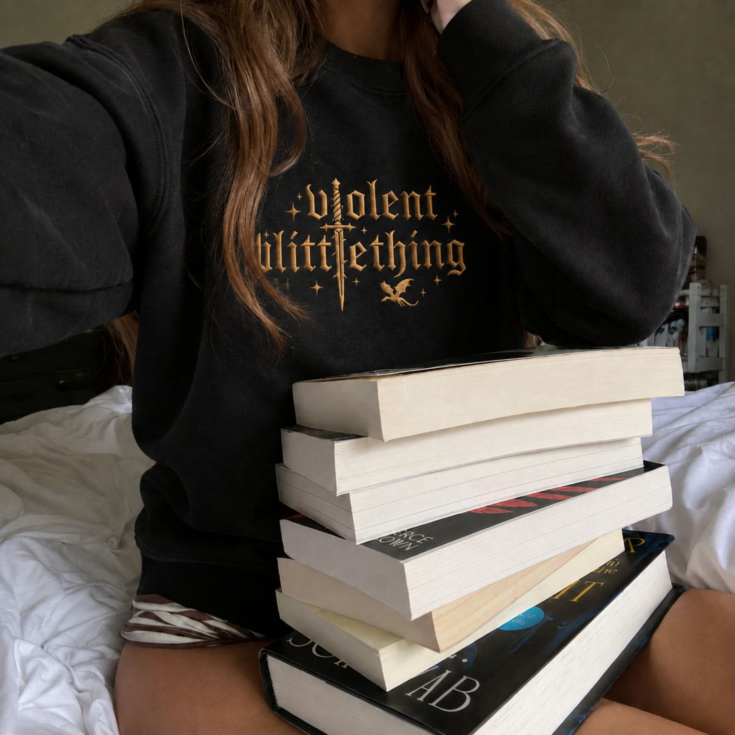 A person wearing a black sweatshirt with a gold gothic-style graphic reading 'violent little thing' sits on a bed with a tall stack of books in front of them.