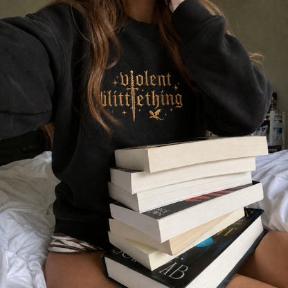 A person wearing a black sweatshirt with a gold gothic-style graphic reading 'violent little thing' sits on a bed with a tall stack of books in front of them.