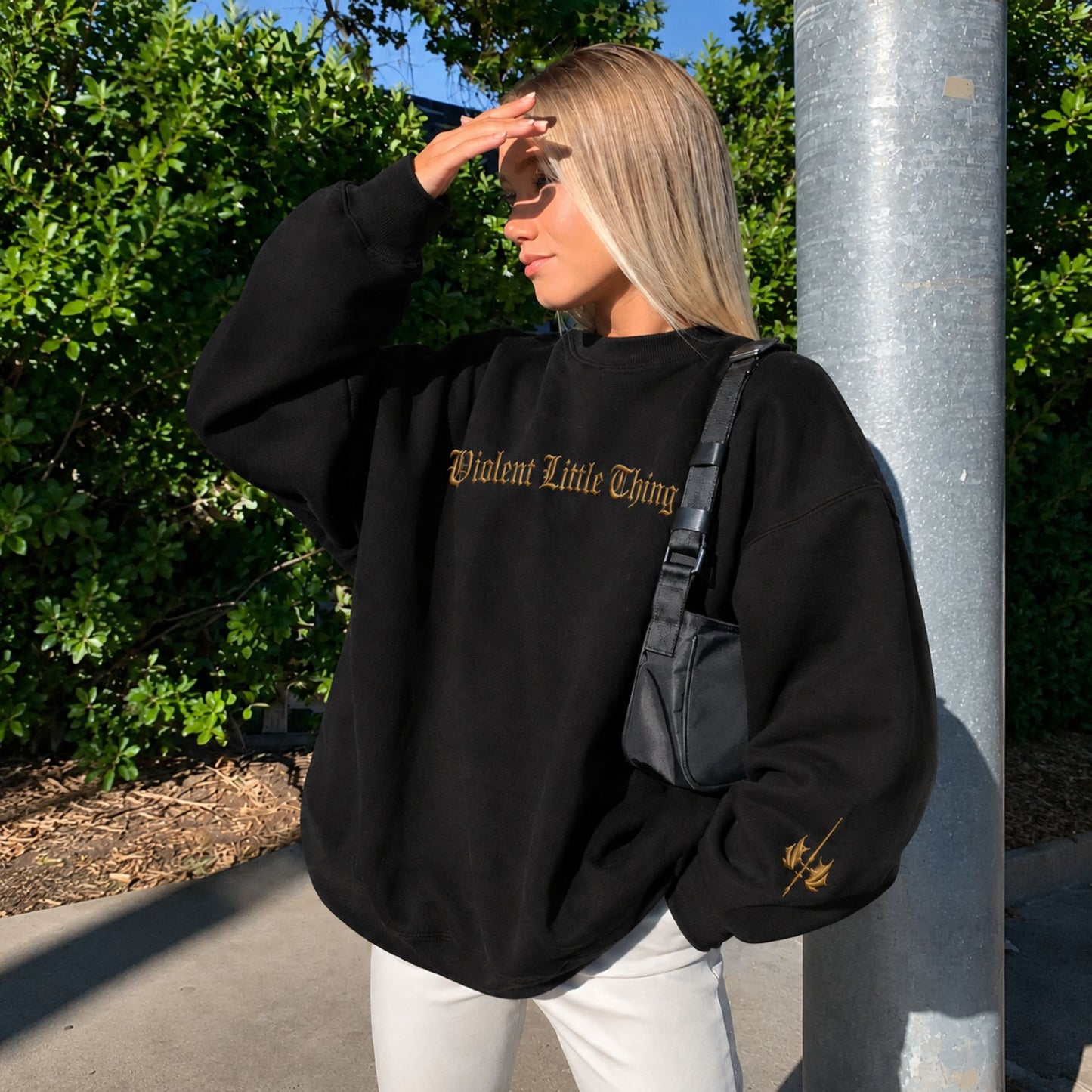 A woman wearing an oversized black sweatshirt with gold Gothic lettering that reads 'Violent Little Thing' on the chest, standing outdoors next to a metal pole with green foliage in the background.