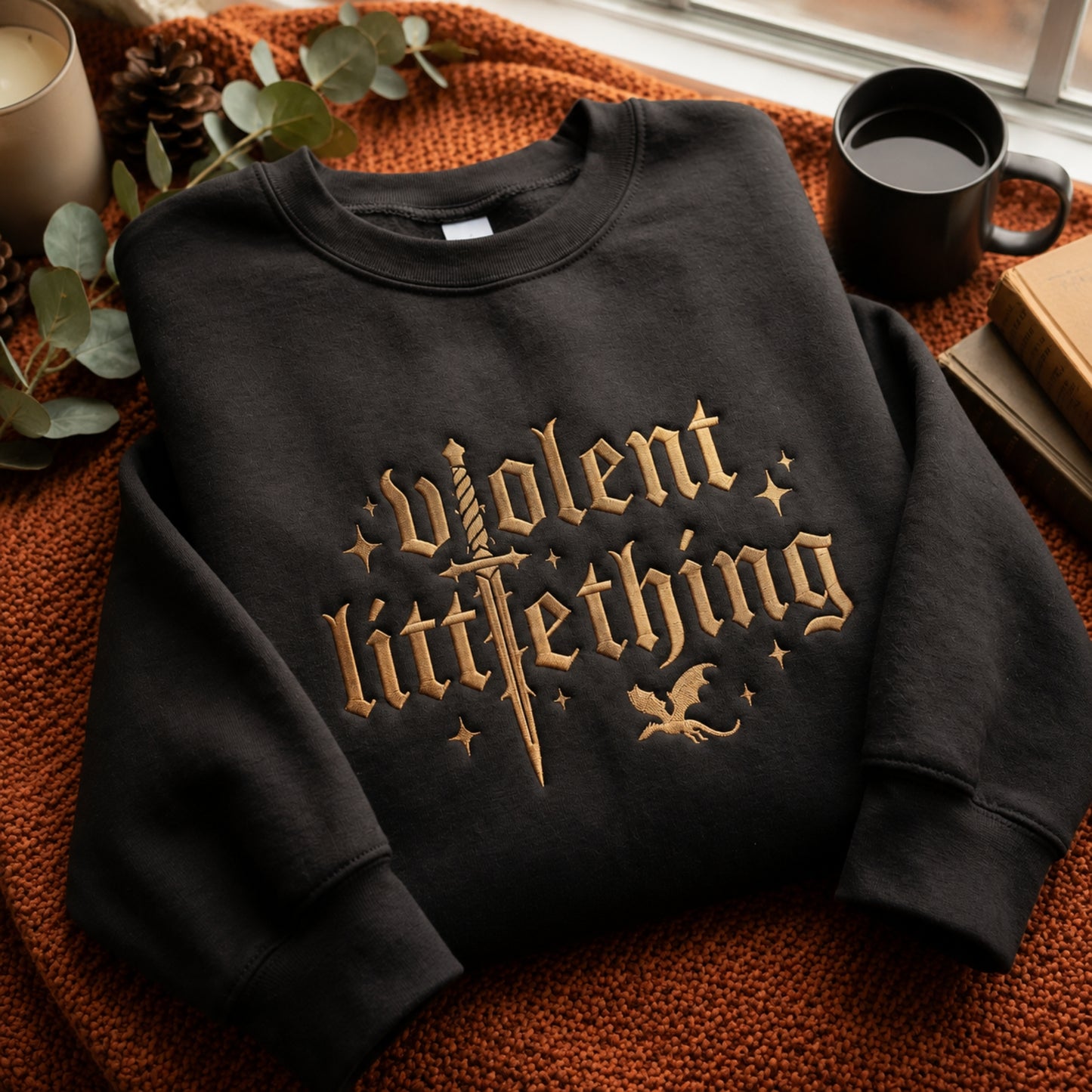 A black crewneck sweatshirt with a gothic-style gold graphic reading 'violent little thing' across the front, styled on an orange knit blanket with a mug, candle, pinecone, and books nearby.