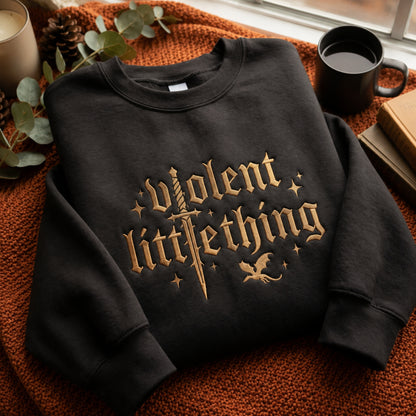 A black crewneck sweatshirt with a gothic-style gold graphic reading 'violent little thing' across the front, styled on an orange knit blanket with a mug, candle, pinecone, and books nearby.
