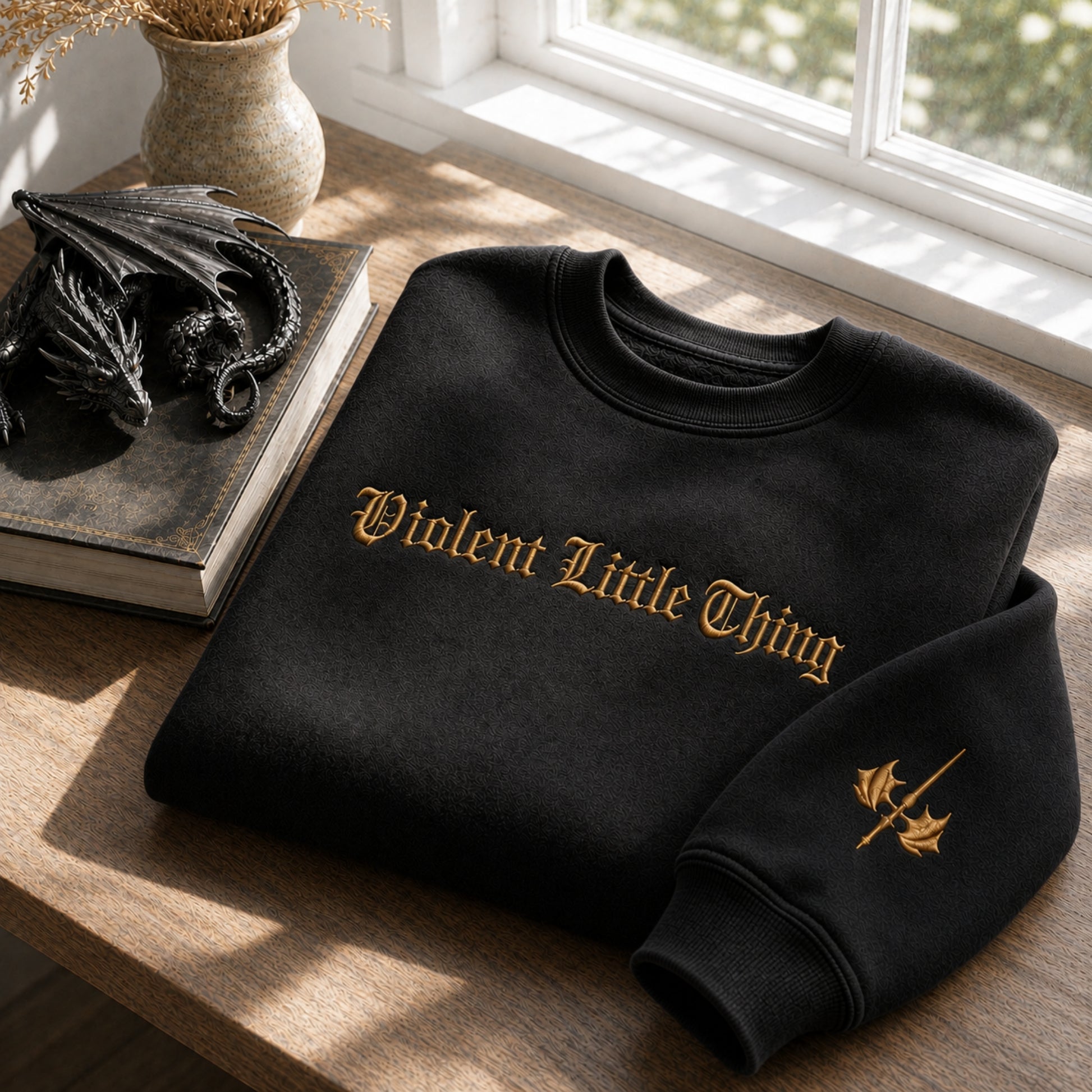 A black crewneck sweatshirt with 'Violent Little Thing' embroidered in gold gothic lettering across the chest, styled on a wooden table next to a dragon figurine and a book.