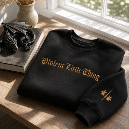 A black crewneck sweatshirt with 'Violent Little Thing' embroidered in gold gothic lettering across the chest, styled on a wooden table next to a dragon figurine and a book.