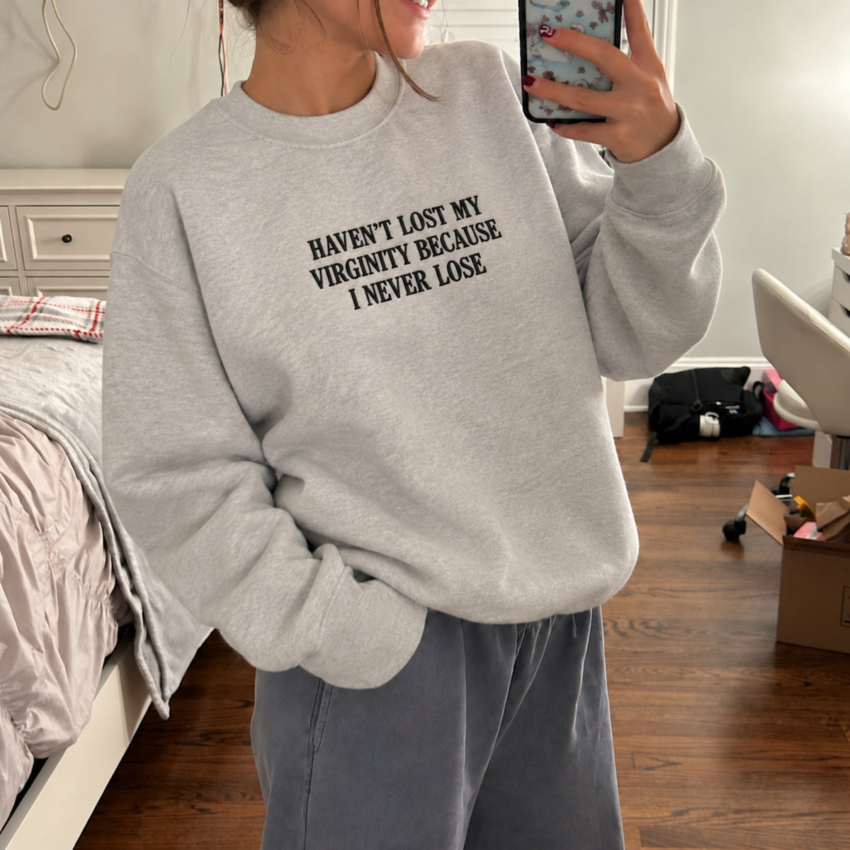 A woman takes a mirror selfie in a bedroom while wearing a light gray crewneck sweatshirt with the black printed text 'HAVEN'T LOST MY VIRGINITY BECAUSE I NEVER LOSE.' and gray sweatpants.
