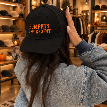 A person wearing a black baseball cap with orange embroidered text reading 'PUMPKIN SPICE GUNT' stands in a boutique store. The person is seen from behind, wearing a light blue denim jacket, with racks of clothing and warm lighting in the background.
