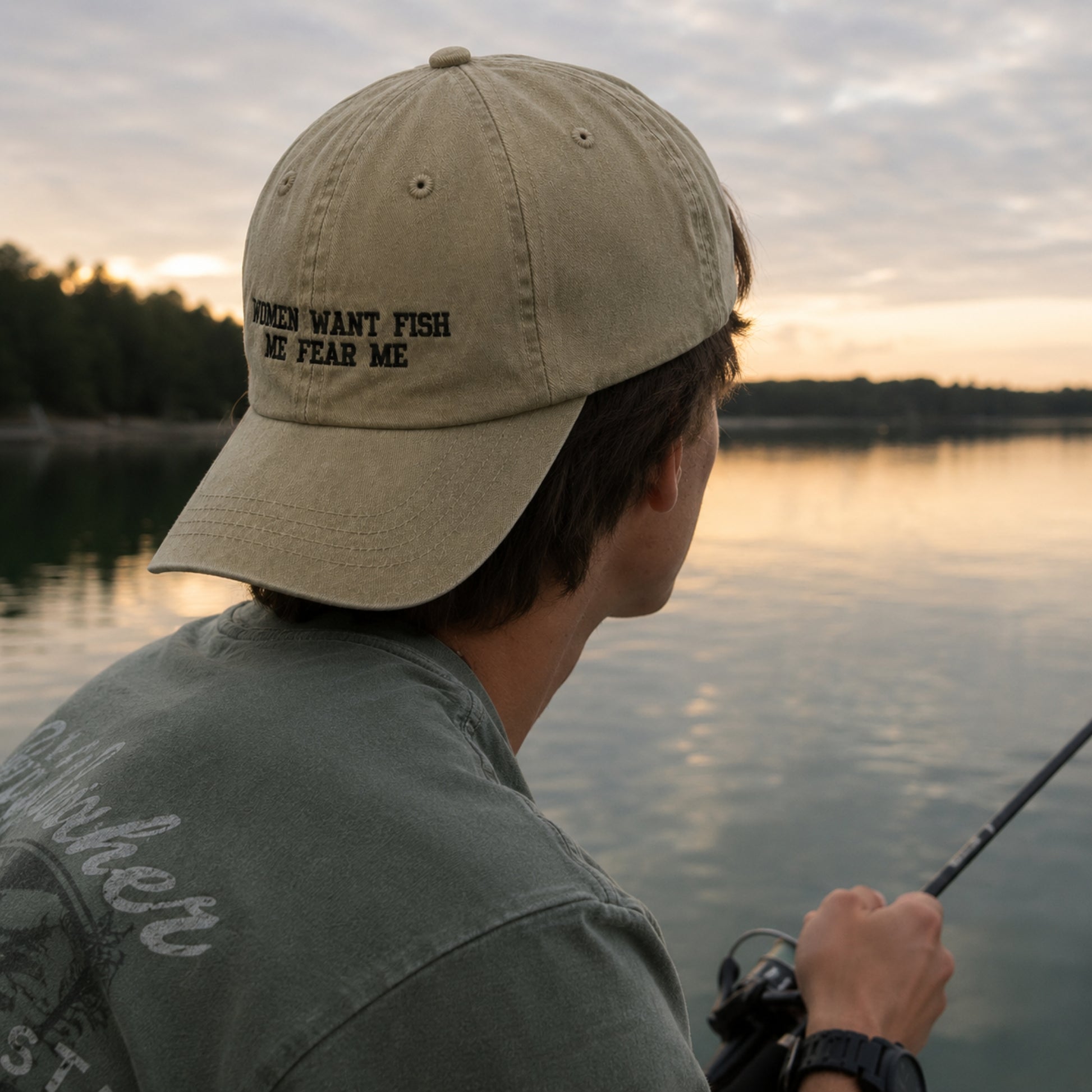 A man wearing a beige baseball cap with the embroidered text 'WHEN I WANT FISH I FEAR ME' in black, shown from behind as he fishes by a lake at sunset.