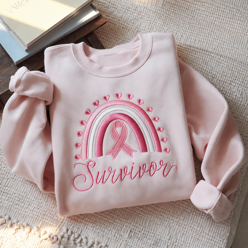 A light pink sweatshirt laid flat on a textured beige fabric surface, featuring an embroidered pink and white rainbow with a pink awareness ribbon and the word 'Survivor' in pink script.