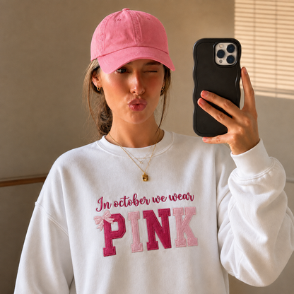 A woman taking a mirror selfie wearing a white sweatshirt with 'In October We Wear PINK' embroidered on the front, a pink baseball cap, and holding a black phone.