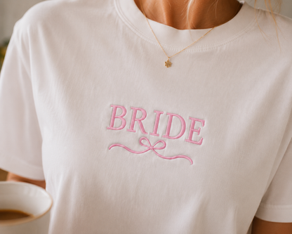 Close-up of a woman wearing a white "BRIDE" embroidered t-shirt with pink script and ribbon design, paired with a delicate gold star necklace — ideal for wedding morning photos, bridal getting-ready outfits, or bride-to-be gift ideas.