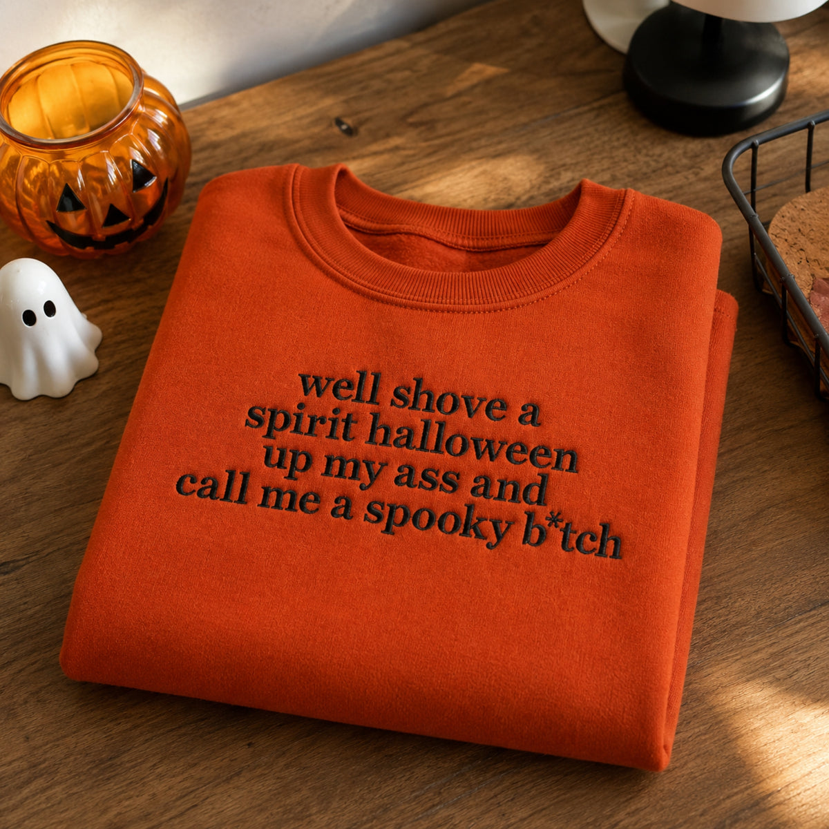 An orange folded crewneck sweatshirt with black embroidered text reading 'well shove a spirit halloween up my ass and call me a spooky b*tch' on a wooden table, styled with a jack-o'-lantern, a small white ghost figurine, and a lamp.